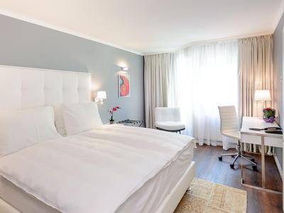 Helles, modernes Hotelzimmer mit Doppelbett, Schreibtisch, Sessel und großen Fenstern mit Vorhängen.