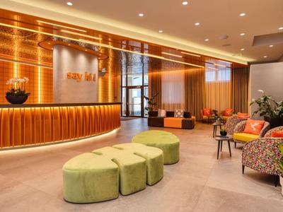 Hall d'hôtel lumineux avec comptoir d'accueil rond, tabourets verts et fauteuils colorés.
