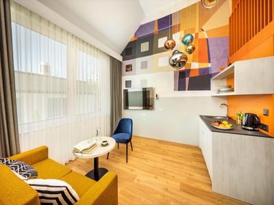 Chambre d'hôtel moderne avec canapé, chaises, kitchenette et grande fenêtre avec rideaux.