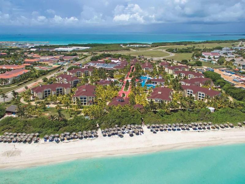 Blick auf einen großen Strandresort mit vielen Gebäuden, Palmen und Sonnenschirmen am türkisfarbenen Meer.