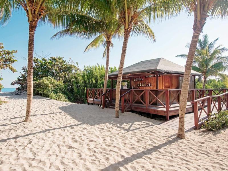 Holzhütte mit Veranda am Sandstrand, umgeben von Palmen und klarem Himmel.