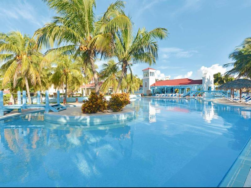 Großer blauer Pool mit Palmen und Sonnenliegen unter klarem Himmel in tropischer Umgebung.