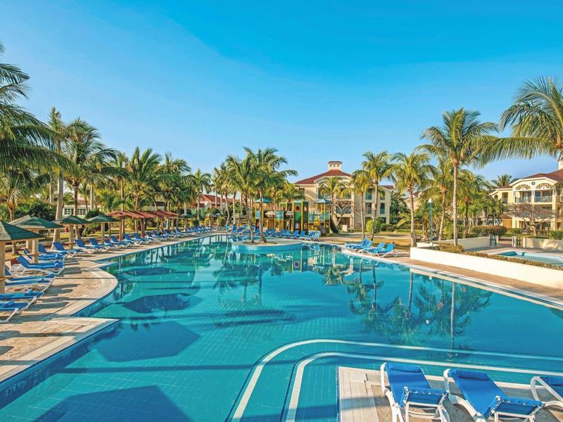 Großer Hotelpool mit blauen Liegestühlen, Palmen und mehrstöckigem Hotelgebäude im Hintergrund.