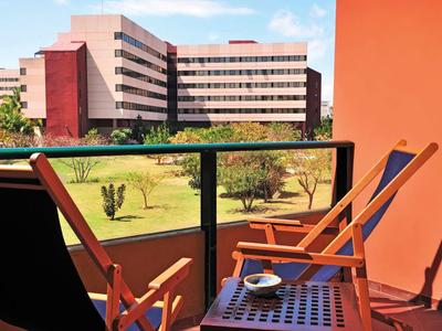 Balkon mit zwei Holzstühlen und Tisch, Blick auf Wiese und modernes Bürogebäude im Sonnenlicht.