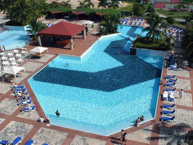 Großer, polygonaler Pool mit blauen Liegestühlen, Sonnenschirmen und Dachterrasse in tropischer Umgebung.