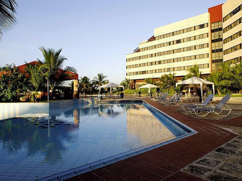 Großer Außenpool mit Liegestühlen vor einem modernen Hotelgebäude bei klarem Himmel.