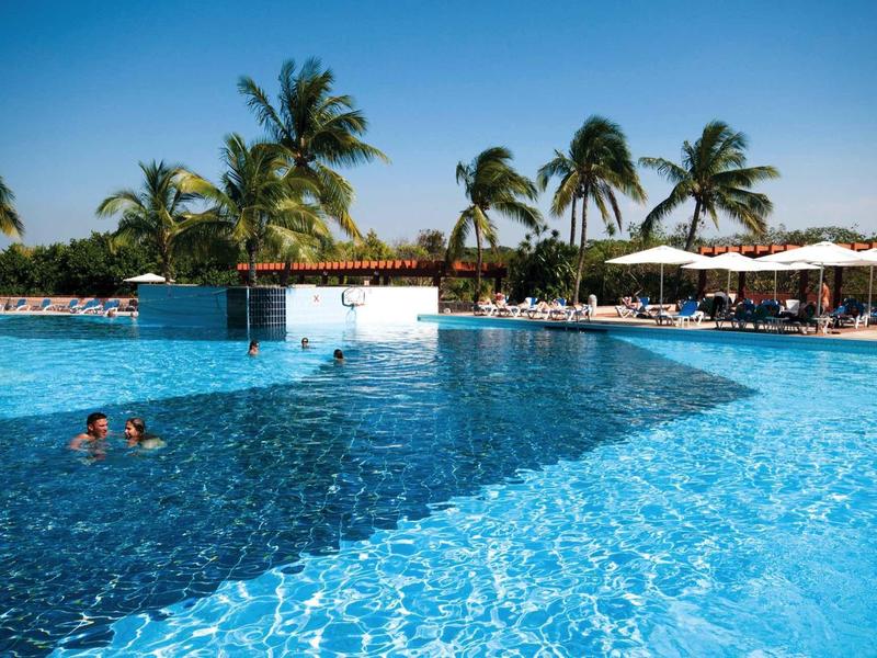 Großer blauer Pool mit Palmen, Sonnenschirmen und Menschen im sonnigen Urlaubsort.