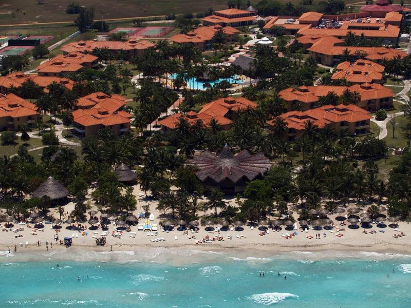 Luftaufnahme eines Resorts mit roten Dächern, grünen Bäumen und einem überfüllten Sandstrand am türkisfarbenen Meer.