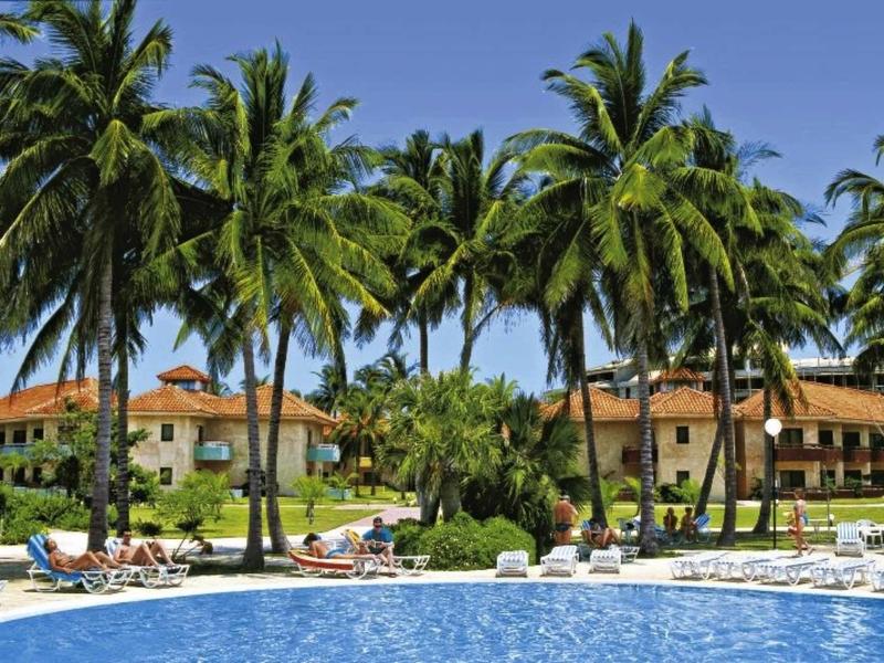 Großer blauer Pool mit Palmen und weißen Liegestühlen vor hellbraunen Hotelgebäuden unter klarem Himmel.