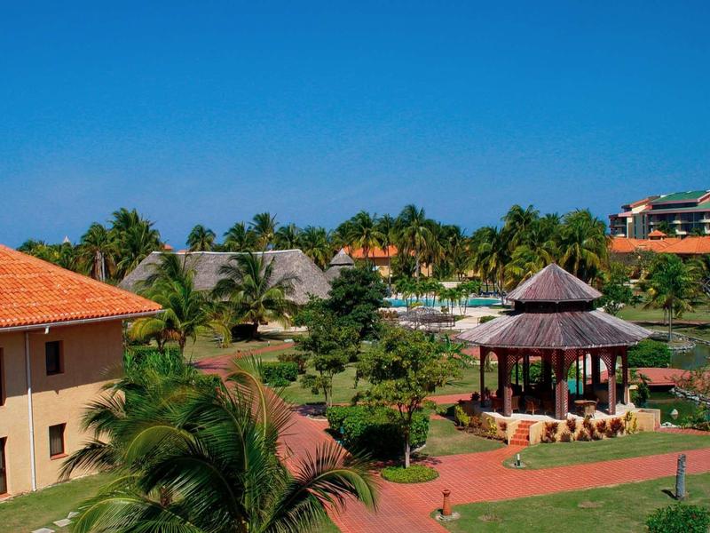 Grüne Gartenlandschaft mit Pavillon, roten Dächern, Palmen und klarem blauem Himmel.
