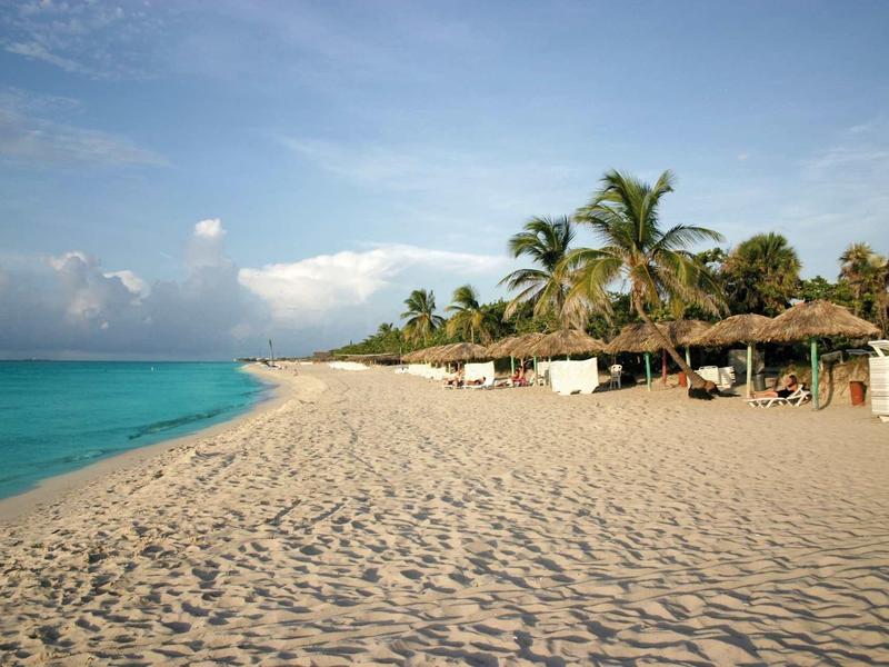 Breiter Sandstrand mit Palmen und Strohhütten am türkisfarbenen Meer unter blauem Himmel.