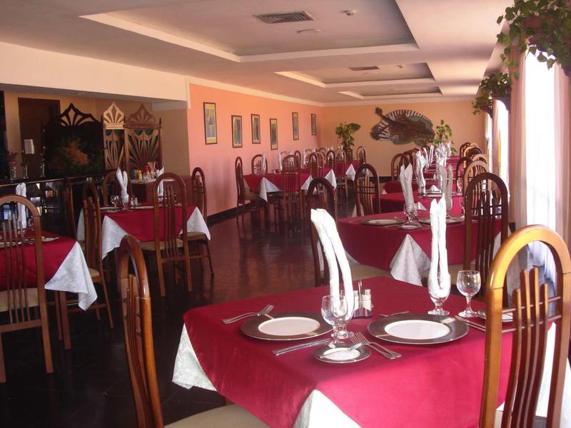 Großer Speisesaal mit Holzstühlen und Tischen, rot-weiße Tischdecken und großen Fenstern links.