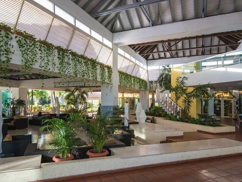 Großer, heller Innenraum mit Pflanzen, weißen Säulen und moderner Lounge mit Treppe und Glaswänden.