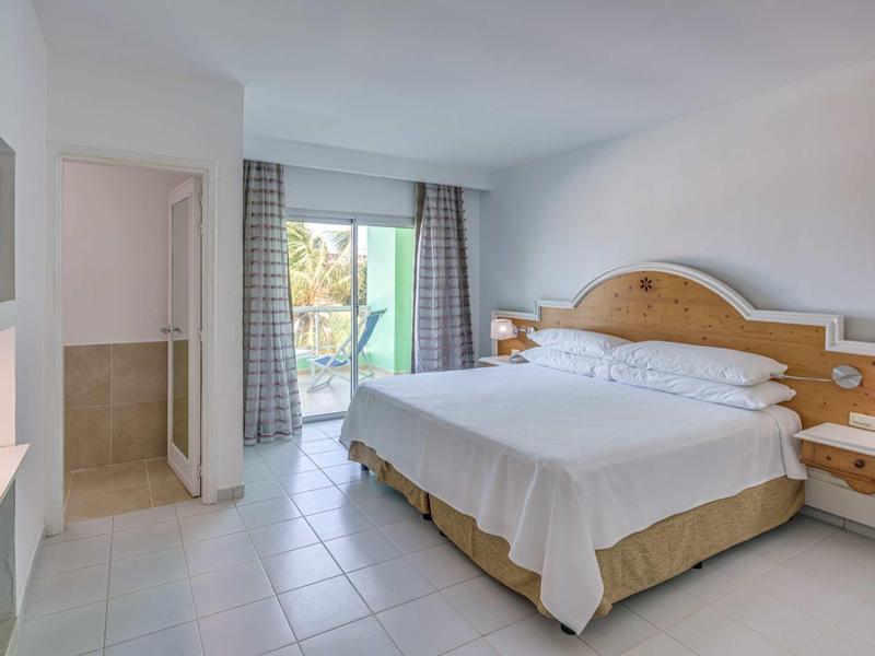 Helles Hotelzimmer mit Doppelbett, Fliesenboden, TV und Balkon mit Blick ins Grüne.