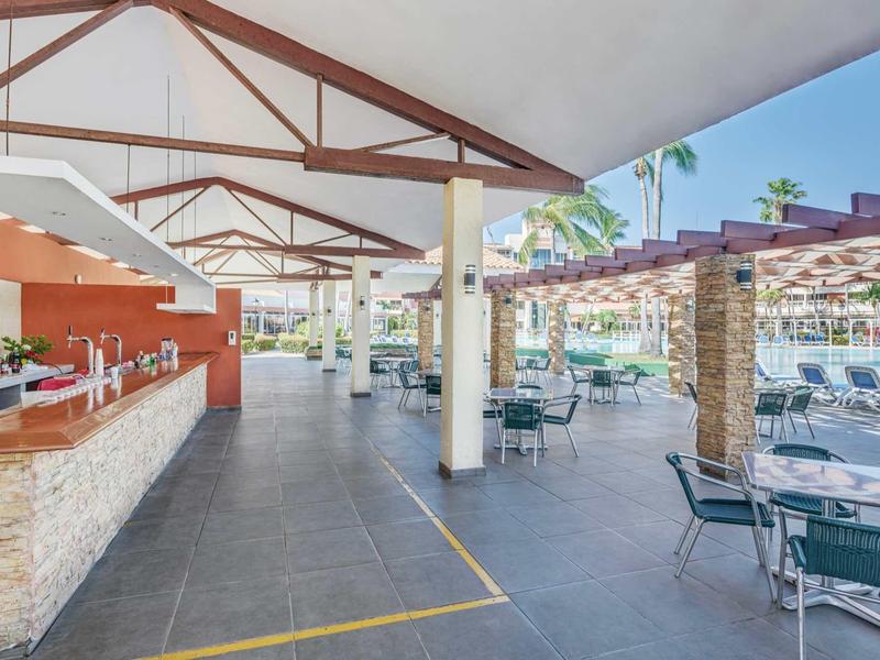 Freiluftbar und Sitzbereich mit Tischen und Stühlen neben einem Hotelpool unter einem überdachten Bereich.