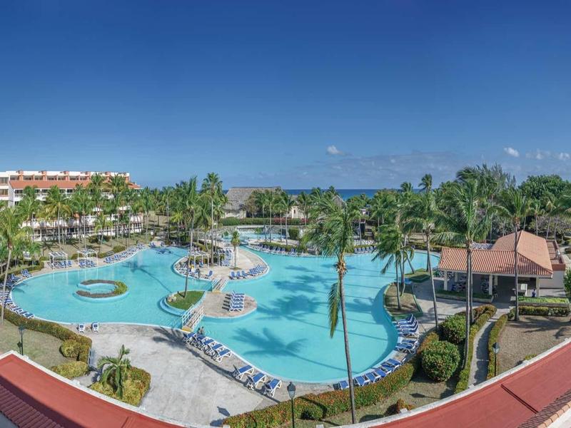 Großer blauer Hotelpool umgeben von Palmen, Liegestühlen und mehrstöckigen Gebäuden unter klarem Himmel.