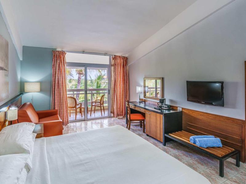 Hell eingerichtetes Hotelzimmer mit Doppelbett, Balkon, orangefarbenen Vorhängen und Fernseher an der Wand.