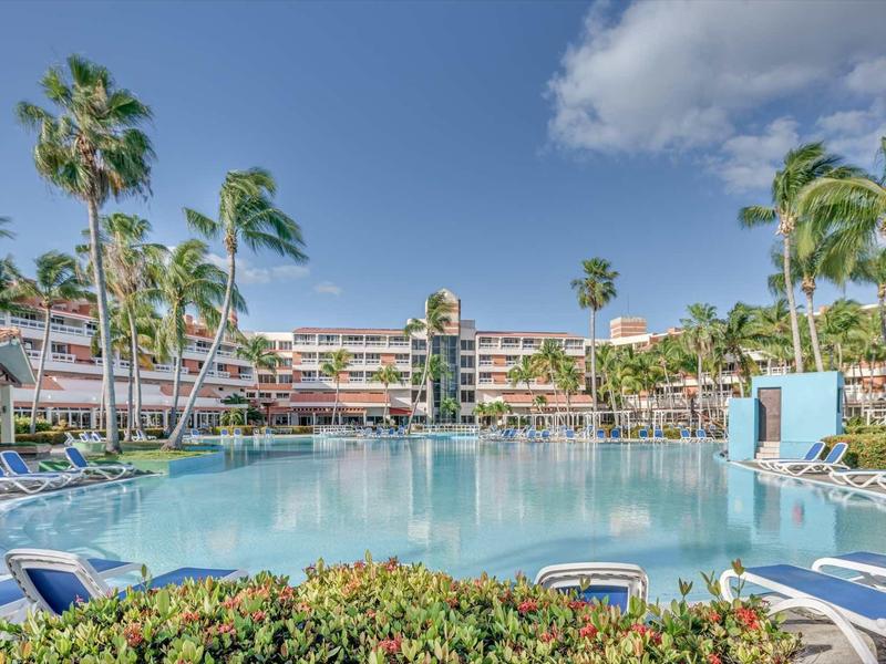 Großer Pool mit Liegestühlen, umgeben von Palmen und mehrstöckigem Hotel unter blauem Himmel.