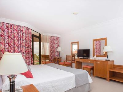 Hell eingerichtetes Hotelzimmer mit Doppelbett, Holzmöbeln und roten Vorhängen am Fenster.