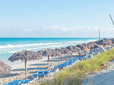 Strand mit blauem Meer, vielen blauen Liegestühlen und Strohdächern unter klarem Himmel.