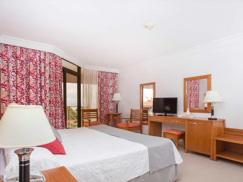 Hell eingerichtetes Hotelzimmer mit Doppelbett, Holzmöbeln und roten Vorhängen am Fenster.