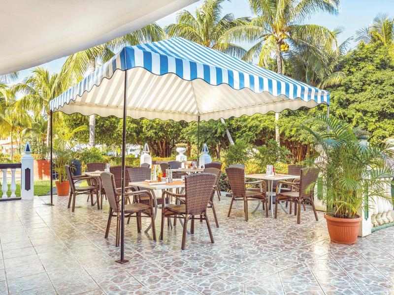 Überdachte Terrasse mit blau-weiß gestreiftem Sonnenschutz, mehreren Tischen und Stühlen, umgeben von Pflanzen.
