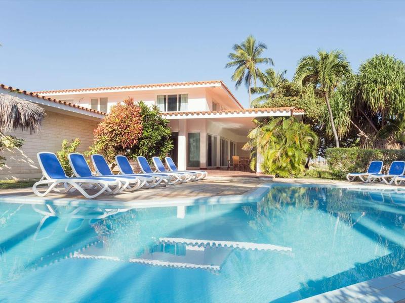 Einladender blauer Pool vor einem Haus mit weißen Liegestühlen und Palmen im Hintergrund.