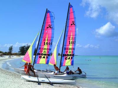 Zwei bunte Segelboote mit Personen am Strand bei blauem Himmel und ruhigem Meer.