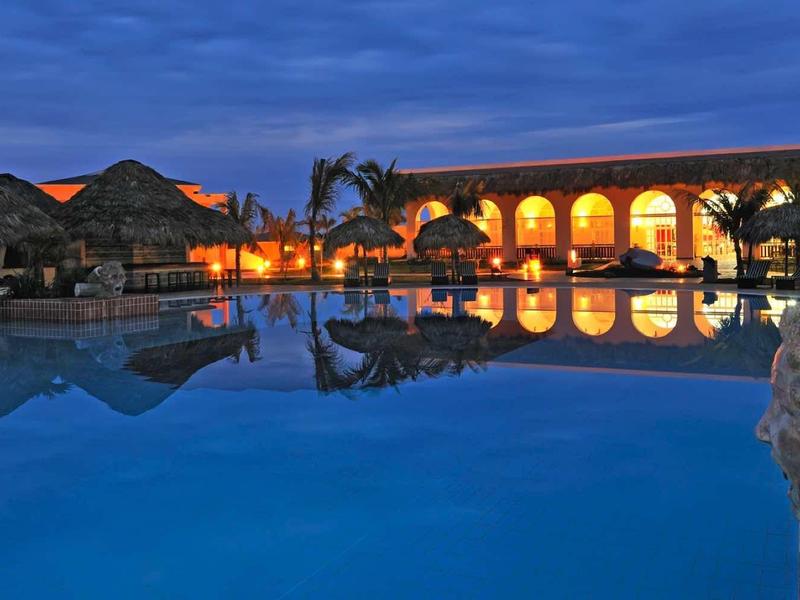 Großer beleuchteter Pool bei Dämmerung mit Spiegelung, Palmen und steinernen Statuen am Rand.