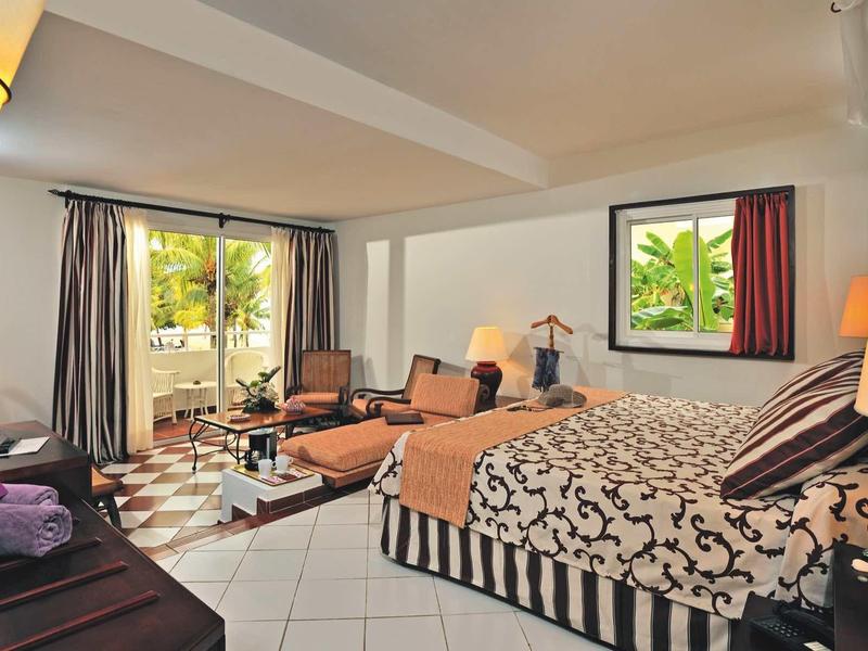 Großes Hotelzimmer mit Doppelbett, Sesseln, Fliesenboden und Blick auf grüne Pflanzen draußen.