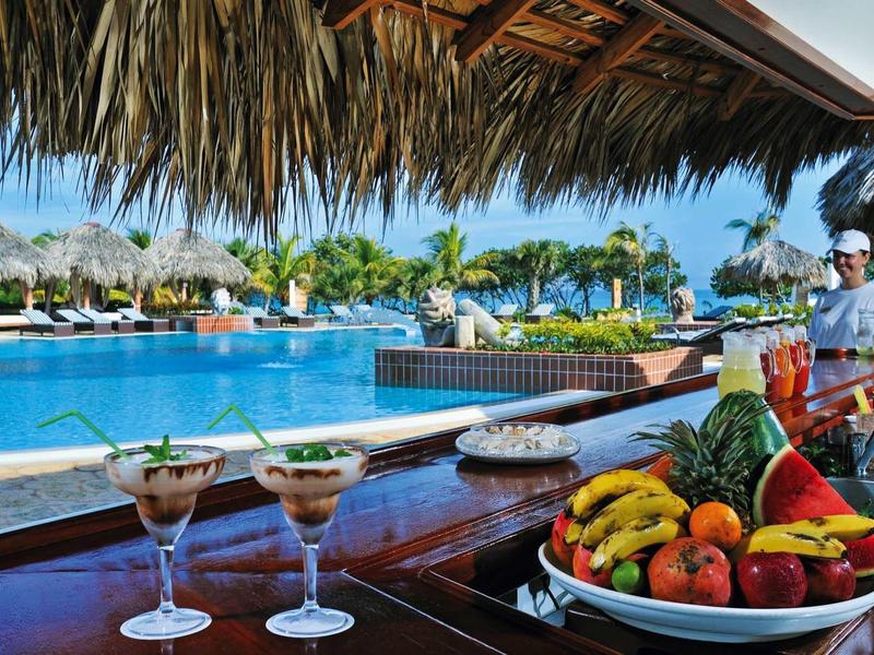 Bar am Pool mit tropischen Früchten, Cocktails und Palmen unter Sonnendach am blauen Wasser.