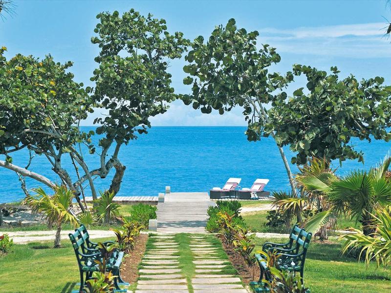 Grüner Gartenweg führt zu Strand mit Liegestühlen und blauem Meer im Hintergrund.