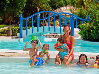 Frau mit vier Kindern im flachen Pool, blauer Bogenbrücke und grünen Bäumen im Hintergrund.
