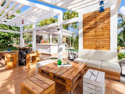 Helle überdachte Outdoor-Sitzbereich mit Holzmöbeln und Blick auf Schwimmbad und Palmen.