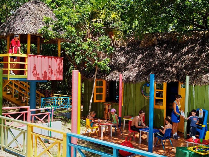 Kinder spielen draußen in einem bunten, mit Palmen gedeckten Spielbereich und Klettergerüsten.