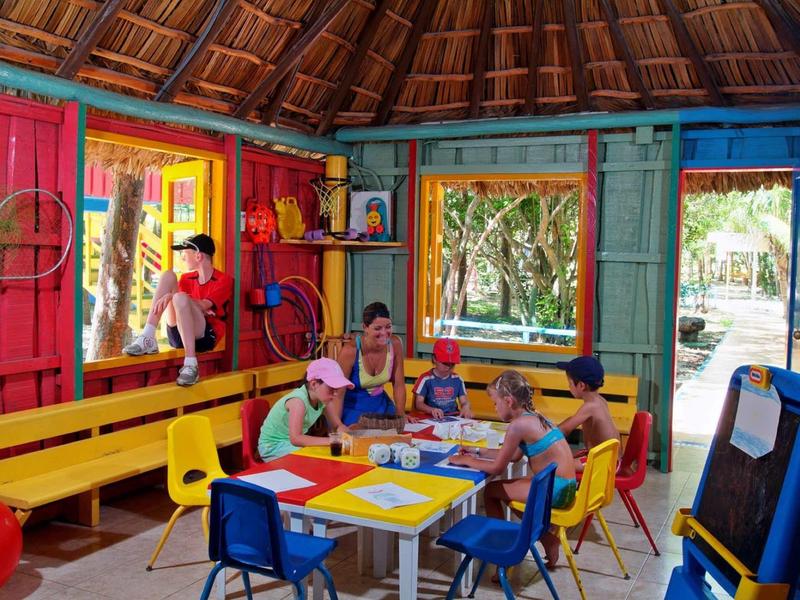 Kinder sitzen an bunten Tischen in einem farbenfrohen, offenen Raum mit Holzdach und Gartenblick.