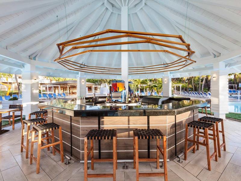Offene Bar mit hölzernen Hockern, großer Deckenbeleuchtung und Blick auf Wasser und Palmen.