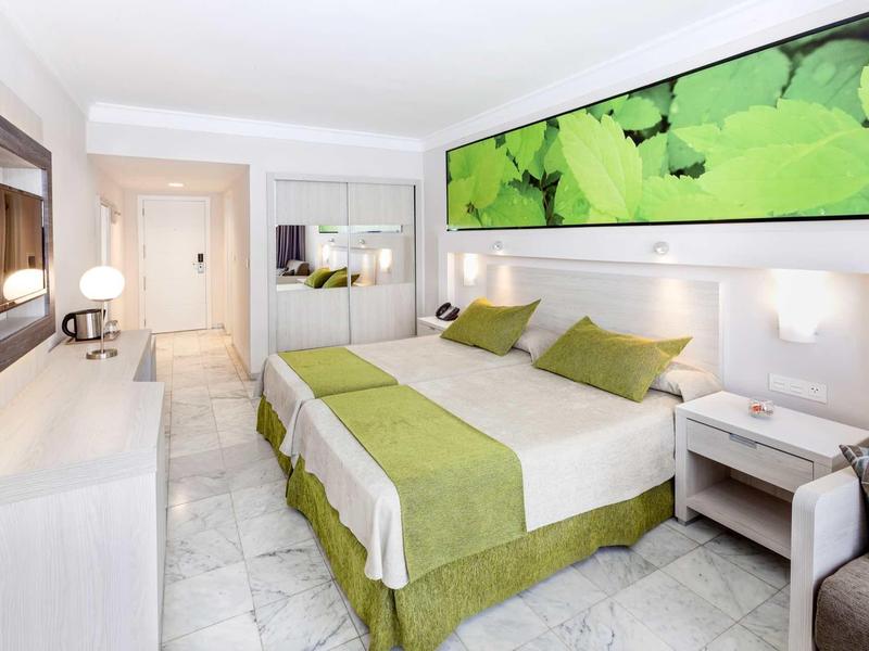 Helles Hotelzimmer mit Doppelbett, grün-weißer Bettwäsche und großem Pflanzenbild über dem Bett.