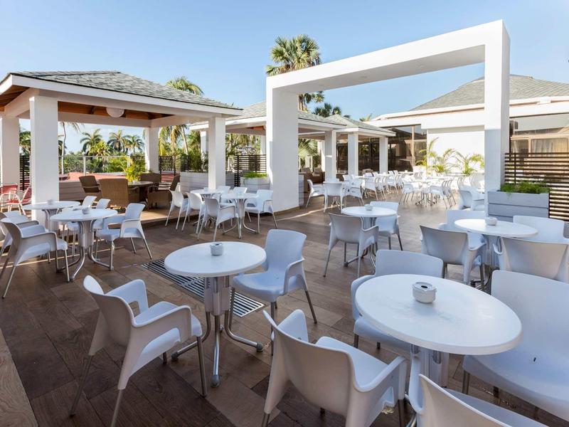 Moderne Terrasse mit weißen Tischen und Stühlen, sonnig, Palmen im Hintergrund, offene Struktur.