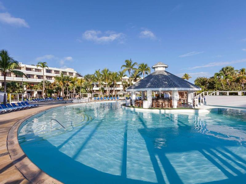Großer Pool mit klarem Wasser, Palmen und blauem Himmel, umgeben von Sonnenliegen und Gebäuden.