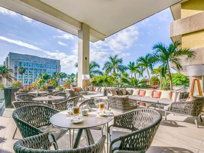 Moderne Terrasse mit Korbstühlen, Tischen und Sofas, umgeben von Palmen und urbaner Kulisse.