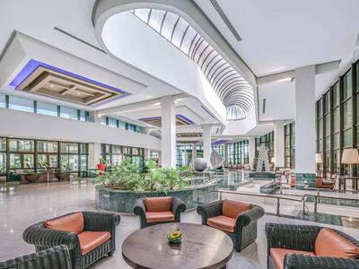 Großer, heller Hotel-Lobbybereich mit Sitzgruppen und Pflanzen unter einer modernen Decke.