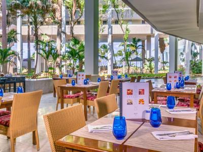 Offener Restaurantbereich mit Holztischen, blauen Gläsern und Blick auf Palmen draußen.