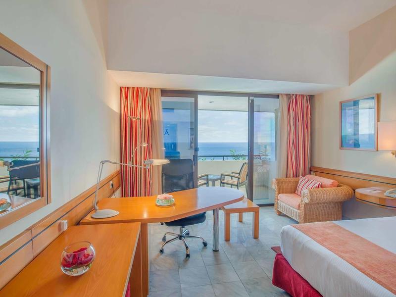 Helles Hotelzimmer mit Meerblick, Schreibtisch, Sofa und Doppelbett.