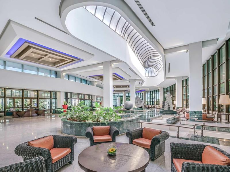 Großer, heller Hotel-Lobbybereich mit Sitzgruppen und Pflanzen unter einer modernen Decke.