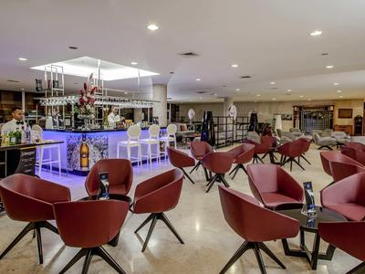 Moderner heller Barraum mit roten Sesseln, gläsernem Bartresen und Deckenbeleuchtung.