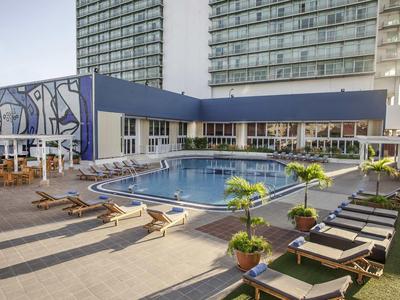 Moderner Außenpool mit Liegestühlen, Pflanzen und großen Glasfassaden neben einem Hotelgebäude.