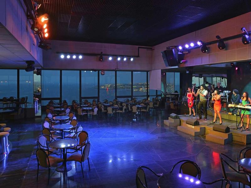 Großer Konzertsaal mit Bühne, Musikern, Sitzplätzen und Panoramafenstern bei Dämmerlicht.