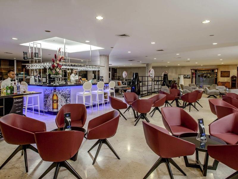 Moderner heller Barraum mit roten Sesseln, gläsernem Bartresen und Deckenbeleuchtung.