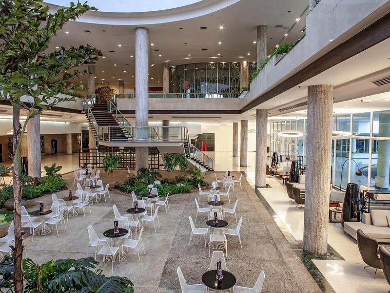 Moderner Innenraum mit Sitzbereich, runden Tischen, Pflanzen und großflächigen Fenstern, helle Farben.